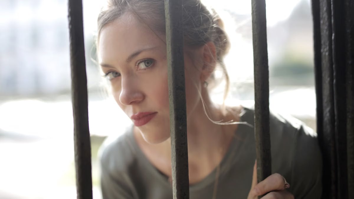 Young beautiful female with bewitching look leaning on iron black bars and looking at camera while standing at fence on sunny day